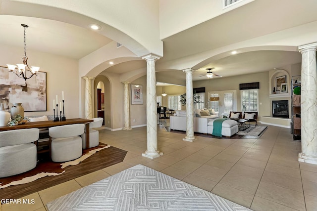 entryway with light tile patterned floors, arched walkways, a large fireplace, ceiling fan with notable chandelier, and ornate columns