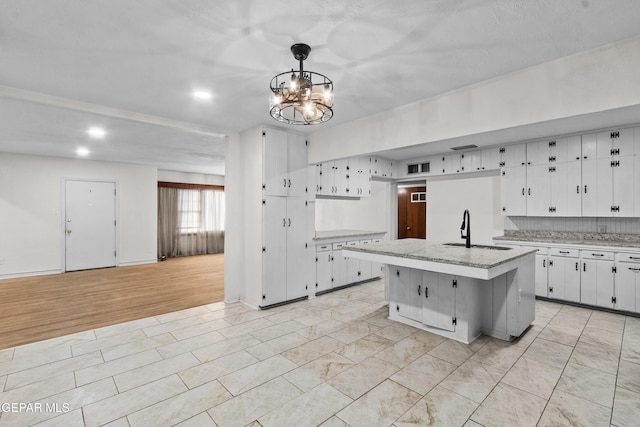 kitchen featuring pendant lighting, a kitchen island with sink, a sink, white cabinets, and an inviting chandelier