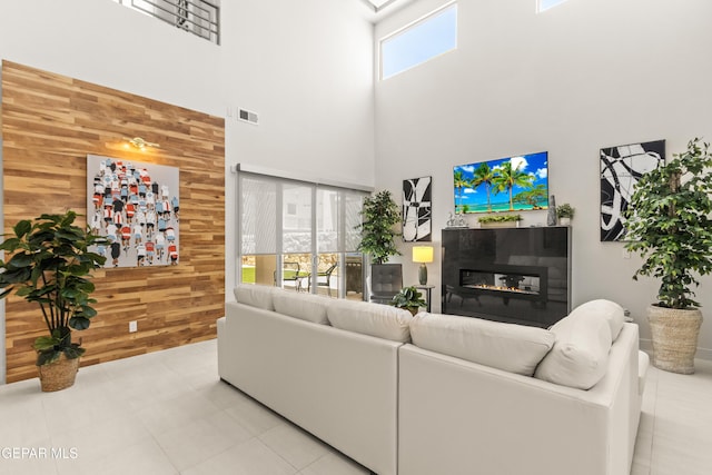 living area featuring light tile patterned flooring, wood walls, a towering ceiling, visible vents, and a glass covered fireplace