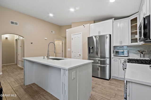 kitchen featuring a center island with sink, visible vents, arched walkways, glass insert cabinets, and appliances with stainless steel finishes