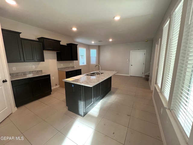 kitchen with light tile patterned floors, an island with sink, a sink, and dark cabinets