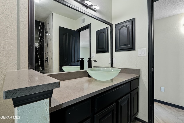 bathroom featuring baseboards, visible vents, a textured wall, walk in shower, and vanity