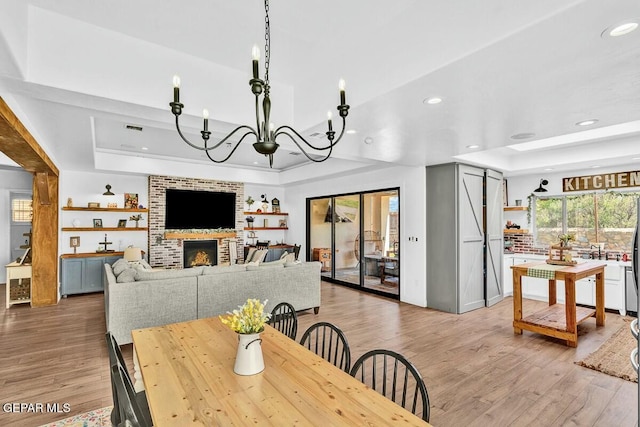 dining room featuring light wood finished floors, a brick fireplace, a raised ceiling, and an inviting chandelier