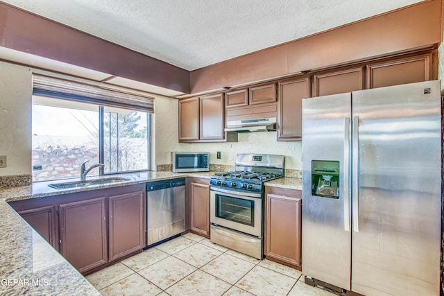 kitchen with a textured ceiling, under cabinet range hood, a sink, appliances with stainless steel finishes, and light stone countertops