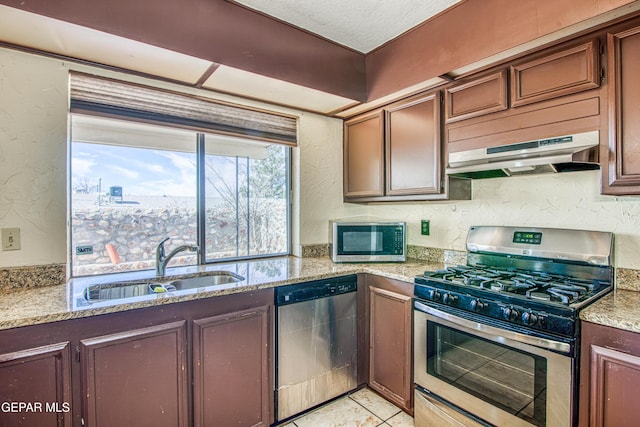 kitchen with a textured wall, light stone counters, under cabinet range hood, stainless steel appliances, and a sink