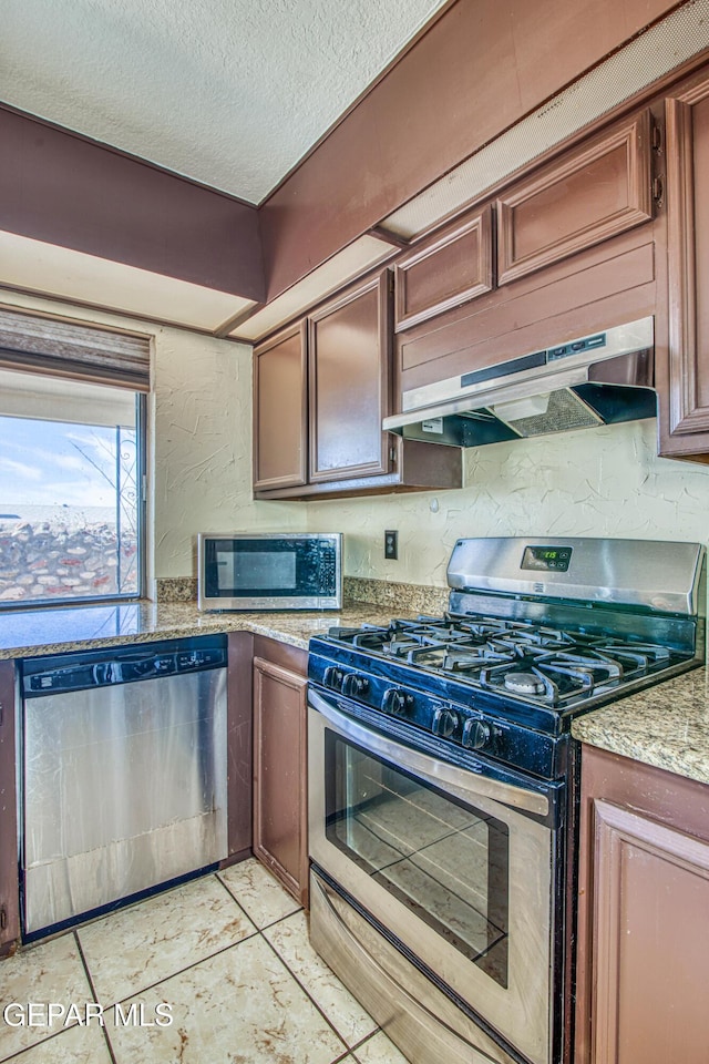 kitchen with a textured ceiling, a textured wall, under cabinet range hood, appliances with stainless steel finishes, and light stone countertops