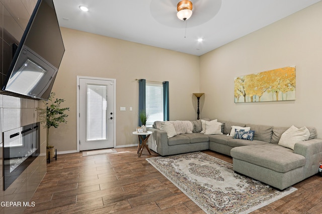 living area with dark wood-type flooring, a tiled fireplace, a ceiling fan, and baseboards