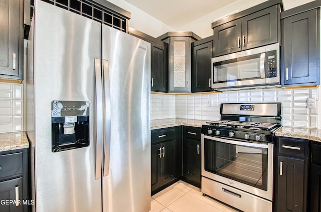 kitchen with light tile patterned floors, decorative backsplash, light stone counters, dark cabinets, and stainless steel appliances