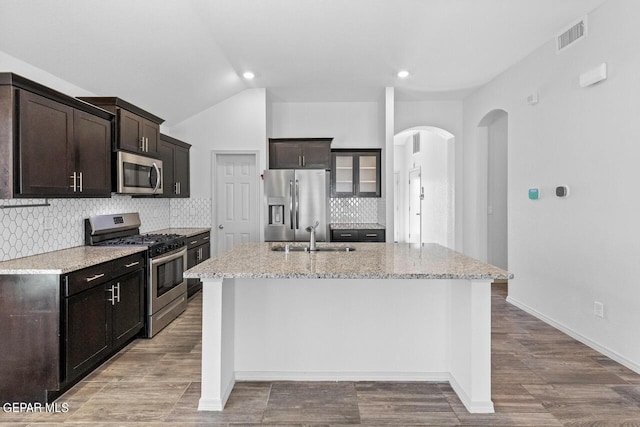 kitchen with arched walkways, appliances with stainless steel finishes, glass insert cabinets, a sink, and an island with sink