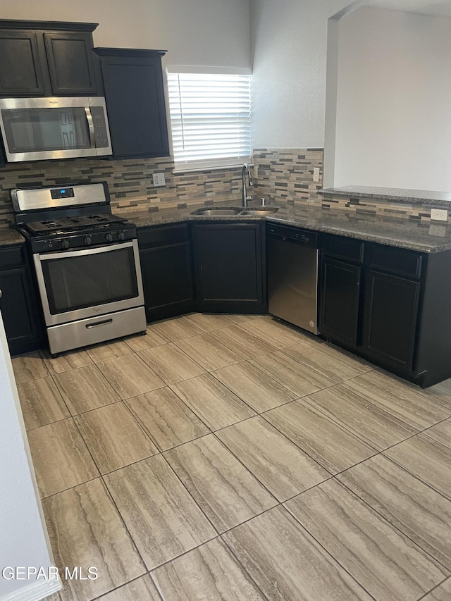 kitchen featuring stainless steel appliances, tasteful backsplash, a sink, dark cabinets, and dark stone counters