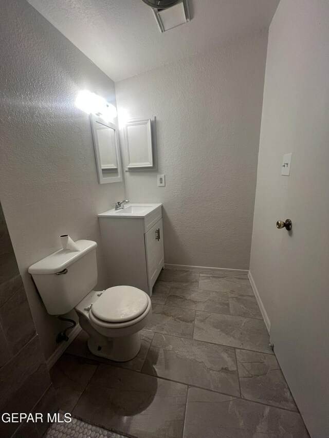 half bath featuring a textured wall, vanity, toilet, marble finish floor, and baseboards