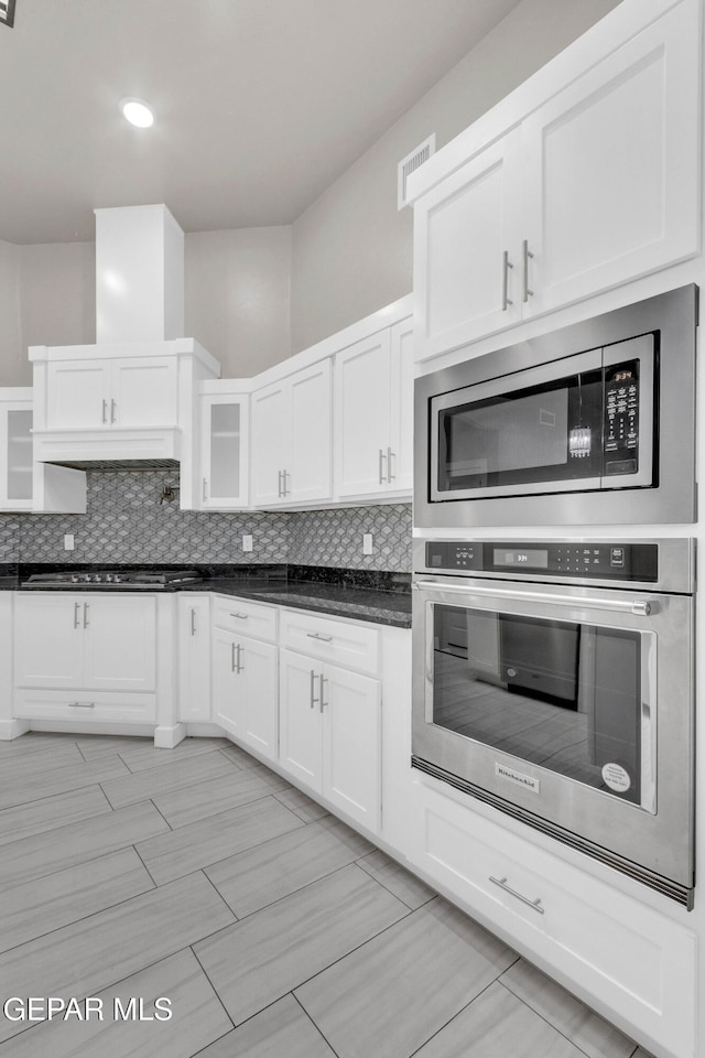 kitchen with white cabinets, appliances with stainless steel finishes, backsplash, dark countertops, and glass insert cabinets