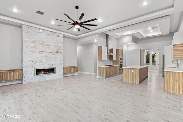unfurnished living room featuring a tray ceiling, a fireplace, visible vents, a ceiling fan, and light wood-type flooring
