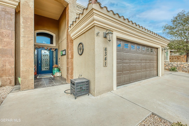 doorway to property with an attached garage, a tile roof, concrete driveway, and stucco siding