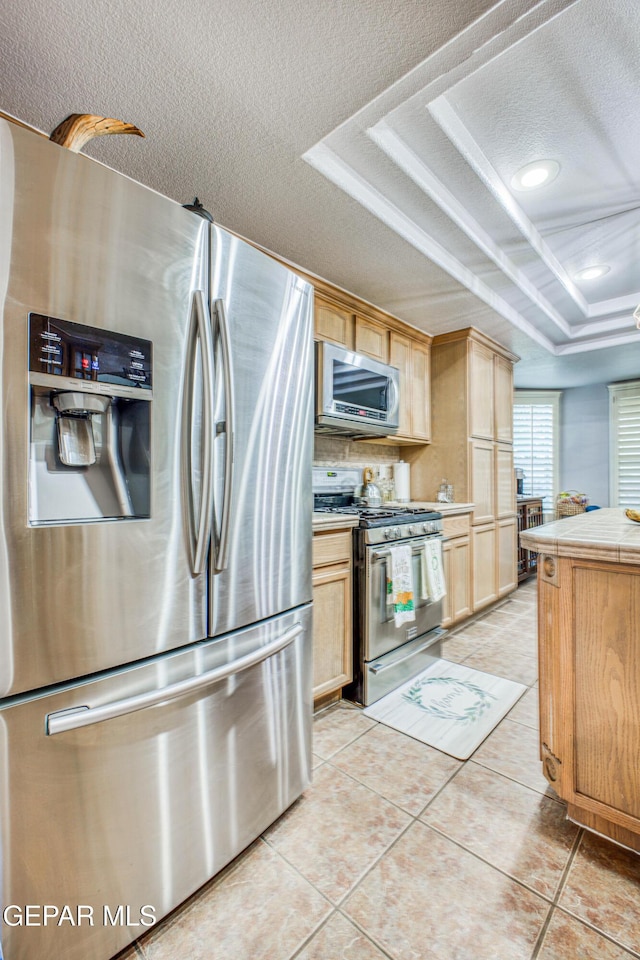 kitchen featuring a textured ceiling, light tile patterned flooring, stainless steel appliances, light brown cabinetry, and a tray ceiling