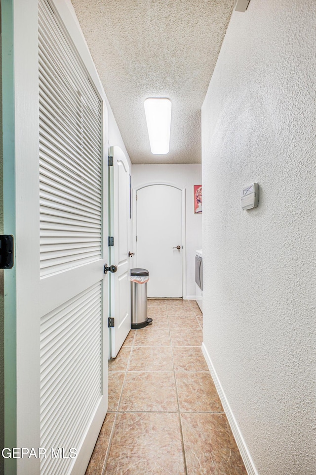 hall featuring a textured wall, a textured ceiling, baseboards, and light tile patterned floors