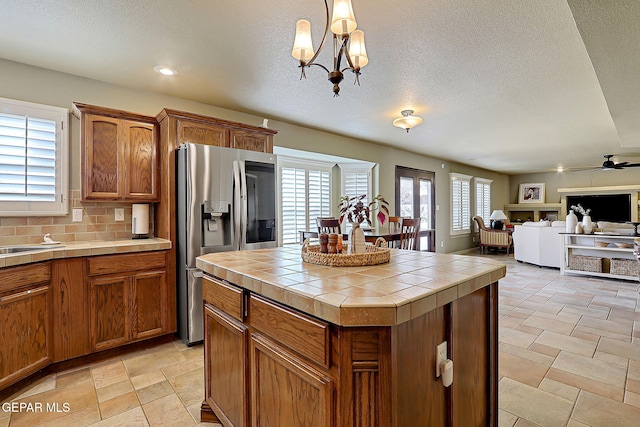 kitchen with stainless steel fridge with ice dispenser, decorative backsplash, tile counters, ceiling fan with notable chandelier, and brown cabinets