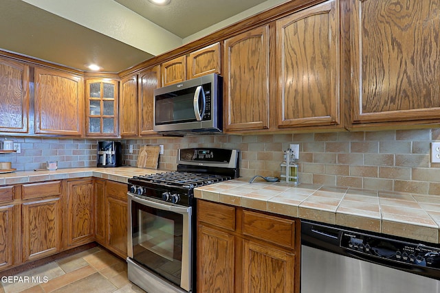 kitchen with brown cabinets, tasteful backsplash, tile countertops, stainless steel appliances, and glass insert cabinets