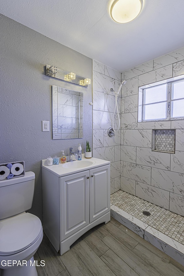 full bath featuring a textured wall, toilet, wood finish floors, vanity, and a tile shower