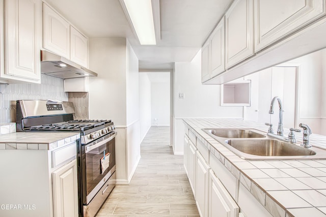 kitchen featuring tile counters, white cabinetry, a sink, stainless steel gas range, and under cabinet range hood