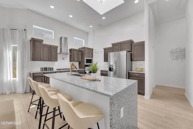 kitchen featuring a skylight, stainless steel appliances, a towering ceiling, a sink, and wall chimney range hood