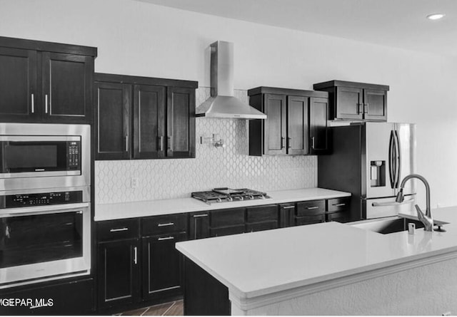 kitchen with stainless steel appliances, a sink, light countertops, dark cabinetry, and wall chimney exhaust hood