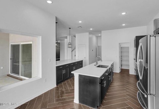 kitchen with light countertops, freestanding refrigerator, a sink, an island with sink, and dark cabinets