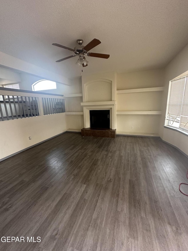 unfurnished living room featuring a textured ceiling, a ceiling fan, a fireplace with raised hearth, and dark wood-style flooring