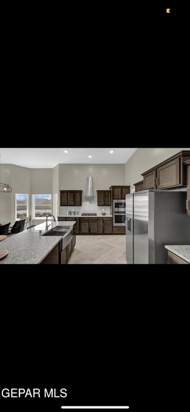 kitchen featuring wall chimney exhaust hood, light stone countertops, stainless steel appliances, dark brown cabinets, and a sink