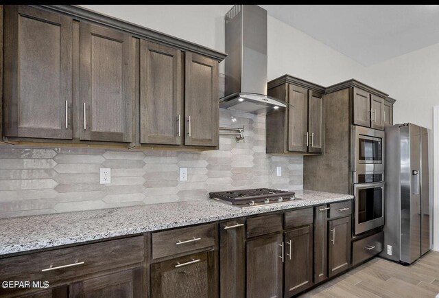 kitchen featuring wall chimney range hood, dark brown cabinets, light stone countertops, stainless steel appliances, and tasteful backsplash
