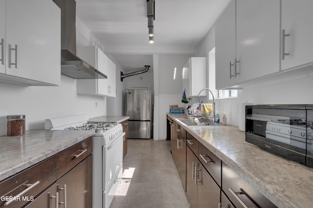 kitchen featuring white gas stove, dark brown cabinetry, a sink, freestanding refrigerator, and wall chimney exhaust hood