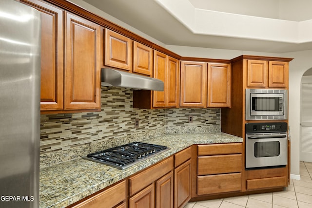 kitchen with stainless steel appliances, decorative backsplash, brown cabinetry, light tile patterned flooring, and under cabinet range hood