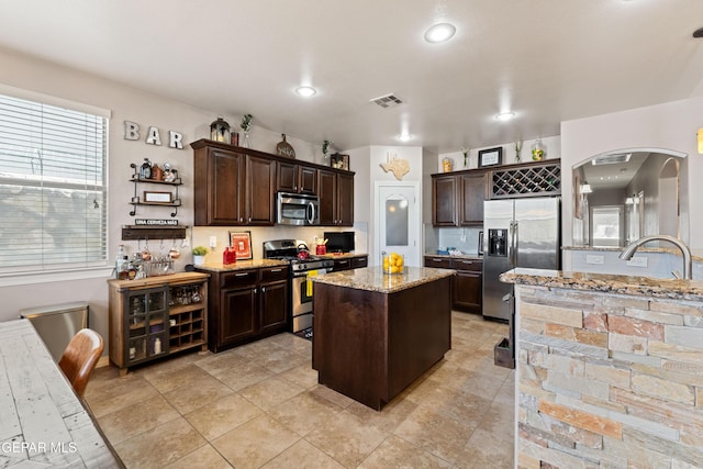 kitchen featuring dark brown cabinetry, a kitchen island, appliances with stainless steel finishes, light stone countertops, and a sink