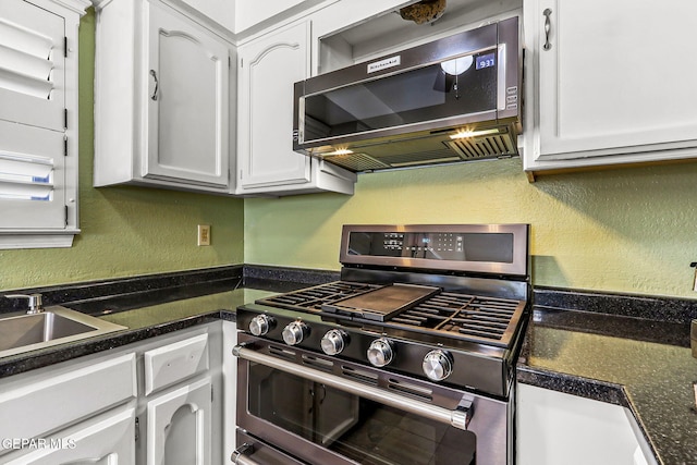 kitchen with stainless steel gas range oven, dark countertops, a textured wall, white cabinets, and a sink