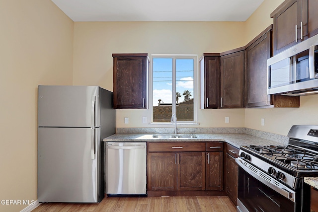 kitchen featuring appliances with stainless steel finishes, light stone countertops, dark brown cabinets, light wood-style floors, and a sink