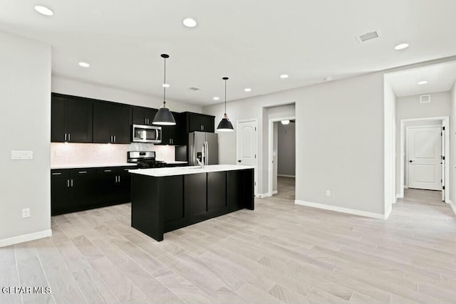 kitchen with dark cabinetry, a center island with sink, light wood finished floors, light countertops, and appliances with stainless steel finishes