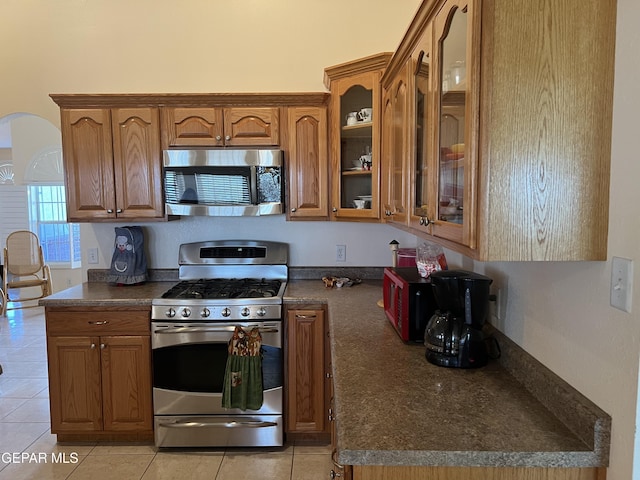 225 Peyton Rd, El Paso, TX 79928 - kitchen featuring stainless steel appliances, dark countertops, brown cabinetry, light tile patterned flooring, and glass insert cabinets