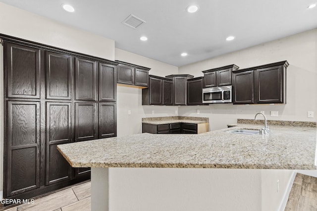 kitchen with light stone counters, dark brown cabinetry, a peninsula, a sink, and stainless steel microwave