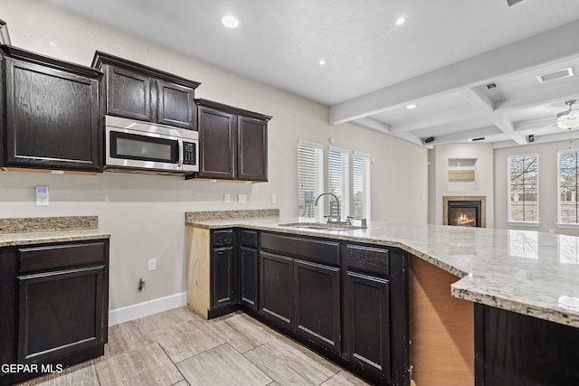 kitchen with a fireplace, stainless steel microwave, a sink, light stone countertops, and beamed ceiling