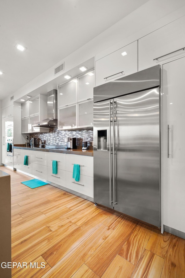 kitchen with visible vents, white cabinets, modern cabinets, stainless steel built in refrigerator, and wall chimney range hood