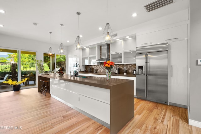 kitchen featuring visible vents, decorative backsplash, wall chimney exhaust hood, stainless steel built in fridge, and white cabinetry