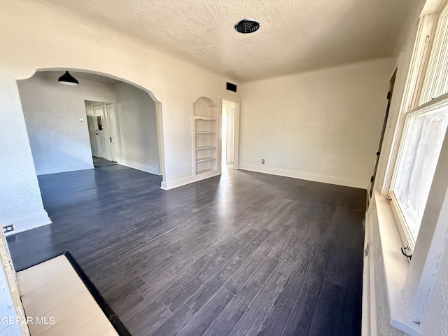 spare room with arched walkways, a textured ceiling, baseboards, built in features, and dark wood finished floors