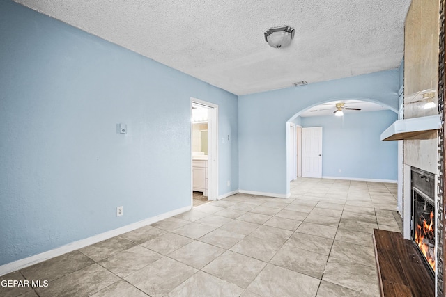 unfurnished living room with a textured ceiling, arched walkways, a fireplace, baseboards, and ceiling fan