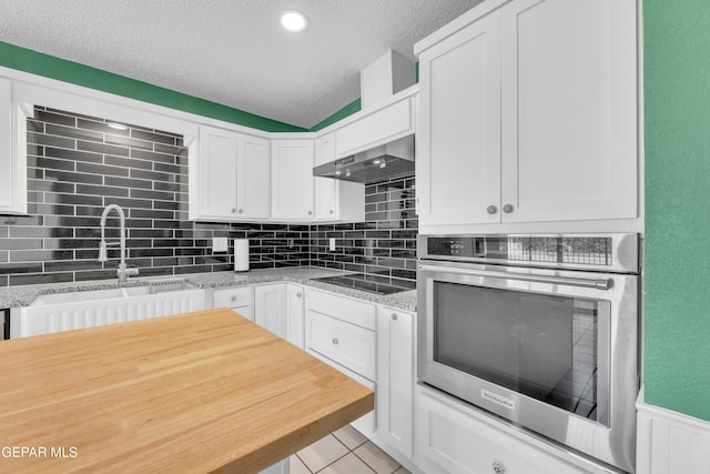 kitchen featuring stainless steel oven, black electric cooktop, wall chimney range hood, white cabinetry, and a sink