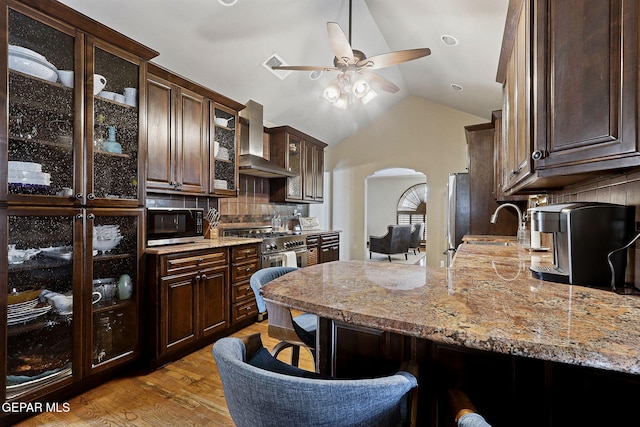 kitchen featuring wall chimney exhaust hood, arched walkways, stainless steel appliances, and dark brown cabinets