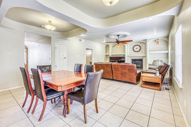 dining space with built in shelves, a glass covered fireplace, light tile patterned floors, a raised ceiling, and ceiling fan