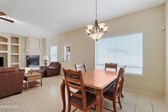 dining room with built in shelves, light tile patterned flooring, a tile fireplace, ceiling fan with notable chandelier, and baseboards