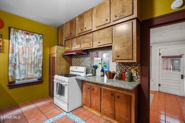 kitchen featuring light tile patterned floors, under cabinet range hood, brown cabinetry, and white range with gas cooktop