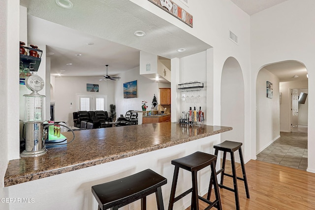 kitchen with ceiling fan, visible vents, a kitchen breakfast bar, light wood-type flooring, and dark countertops