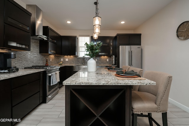 kitchen featuring appliances with stainless steel finishes, wall chimney range hood, dark cabinetry, tasteful backsplash, and pendant lighting